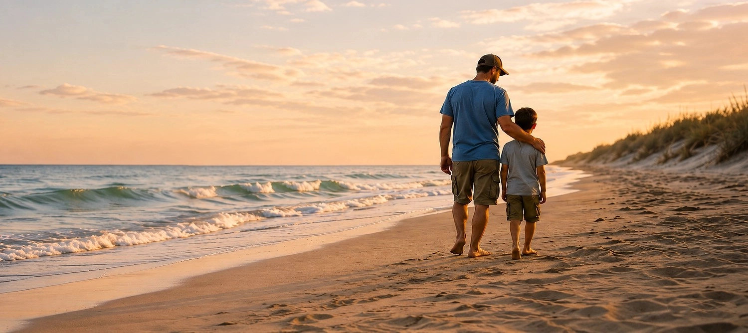 Michael | ChildHeartHealing® - Gesundheits und Hypnose Coach - Ein Vater und sein Sohn spazieren barfuß bei Sonnenuntergang an einem sandigen Meeresstrand entlang, von hinten gefilmt.